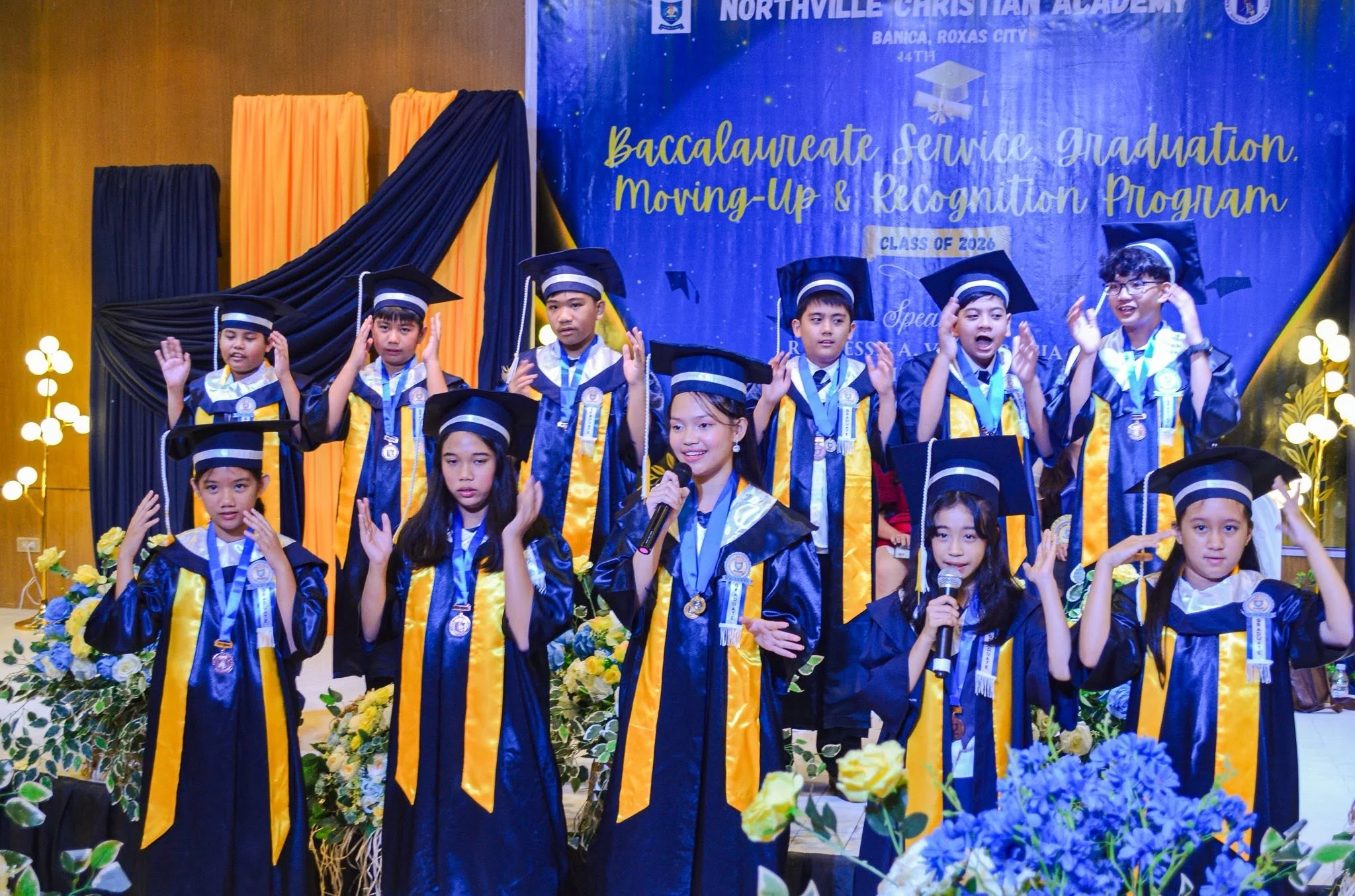 Young children in navy caps and gowns stand on a stage during Northville's Moving Up ceremony, with the school's name on a backdrop behind them.