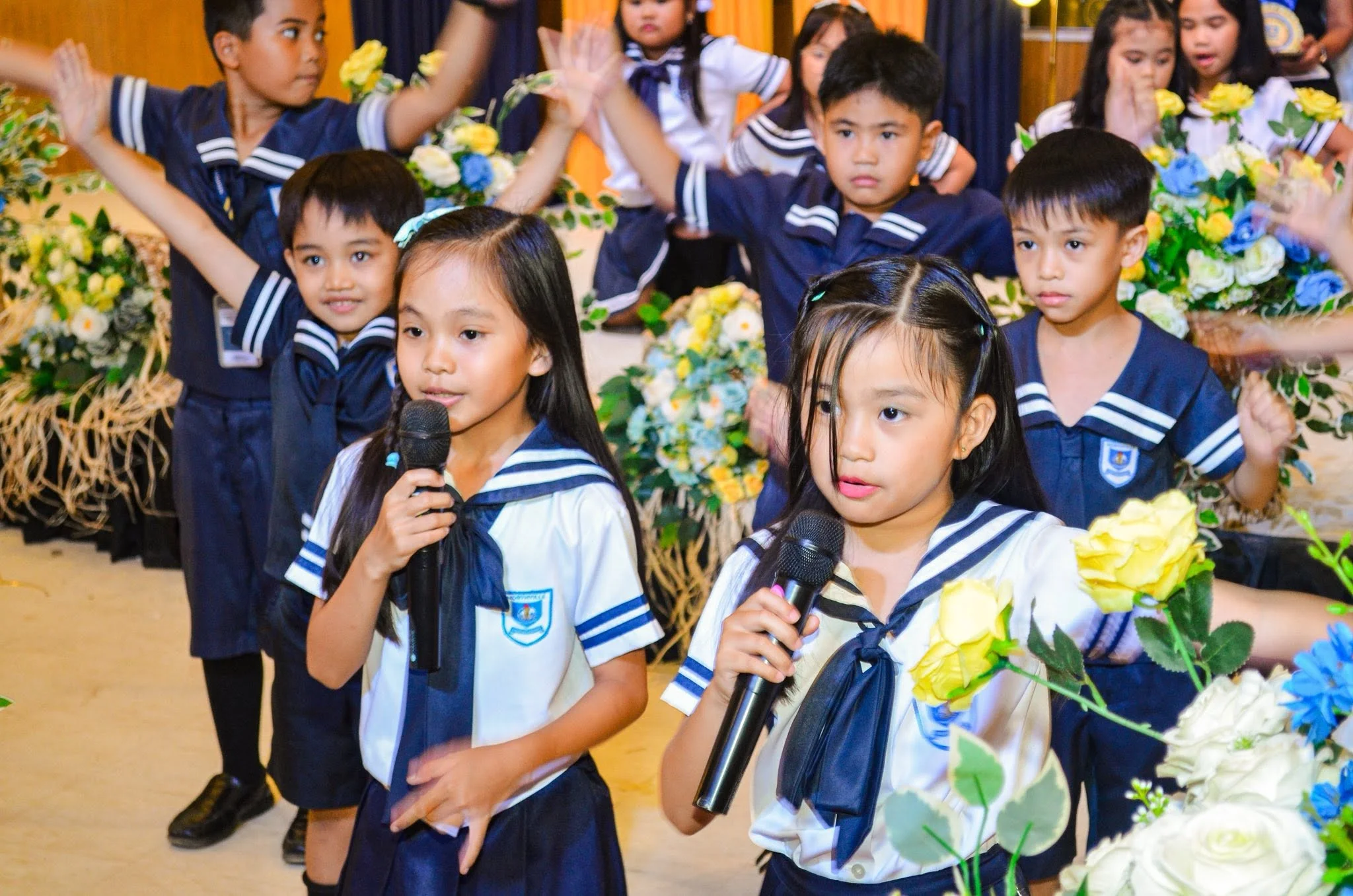 Preschoolers in navy-and-white sailor uniforms sing into microphones on stage, with flowers arranged behind them.