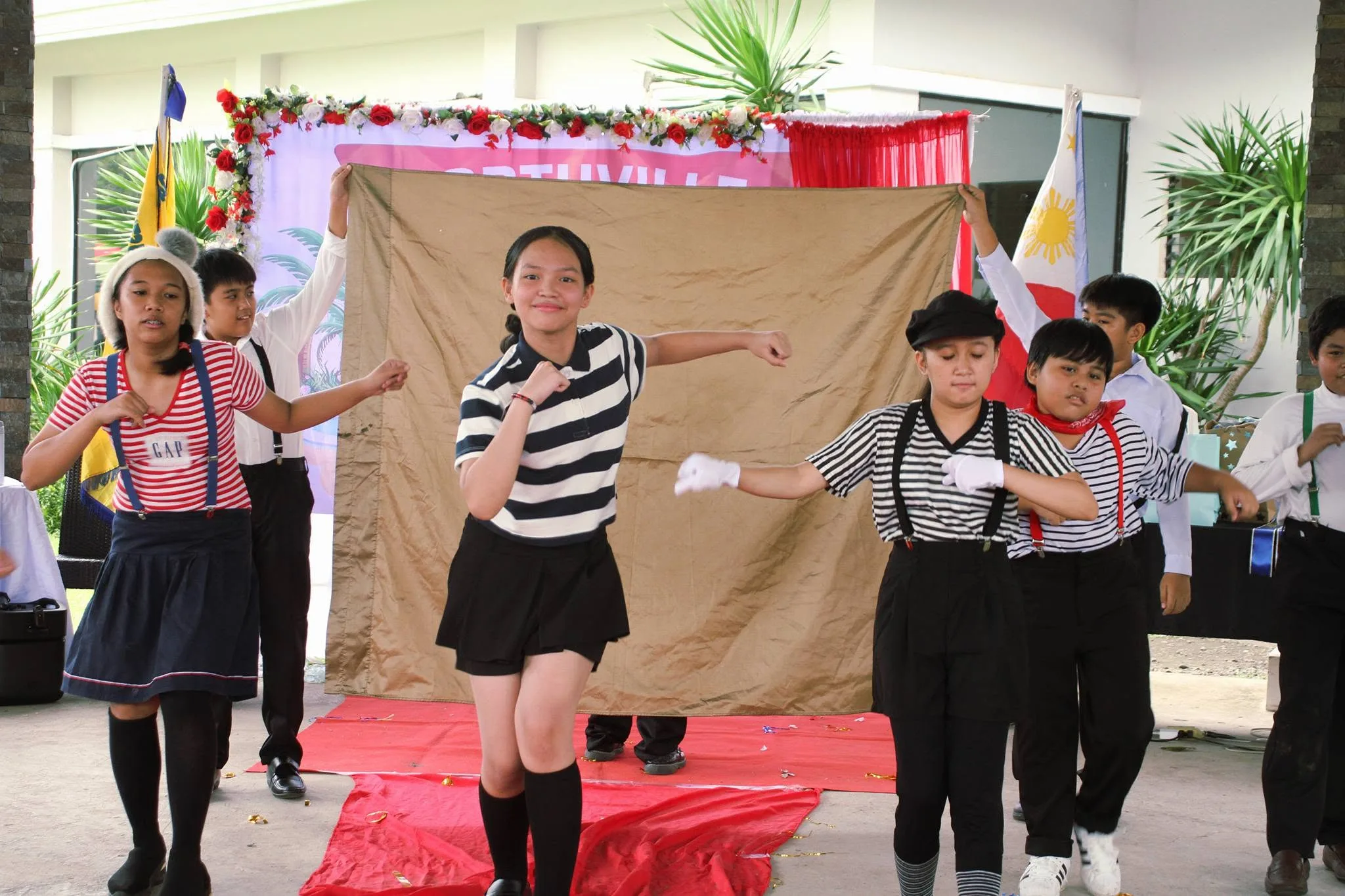 Students in striped shirts and suspenders perform a dance routine in front of a backdrop at a school program.